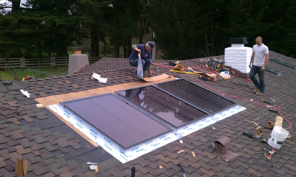 Two workers installing solar panels on a shingled roof surrounded by various tools and equipment.